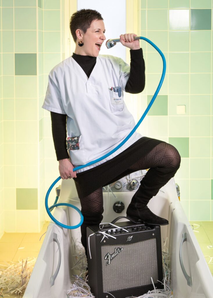 Photographie de Lionel Pralus / Light Motiv, issue de la série intitulée Désordre Ordinaire datant de 2020, représentant une femme debout dans des vestiaires faisant semblant de chanter avec une douchette et une enceinte sous sa jambe relevée, dans le cadre d'un projet tourné autour de mise en scène photographiques avec les patients et professionnels du SAAS.