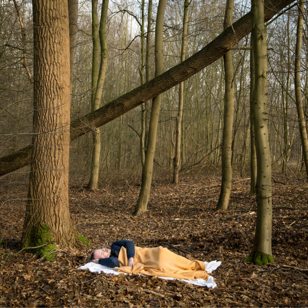 Photographie de Lionel Pralus / Light Motiv, issue de la série intitulée Désordre Ordinaire datant de 2020, représentant un homme allongé sur un drap blanc et recouvert d'une couverture orange dans des bois, dans le cadre d'un projet tourné autour de mise en scène photographiques avec les patients et professionnels du SAAS.