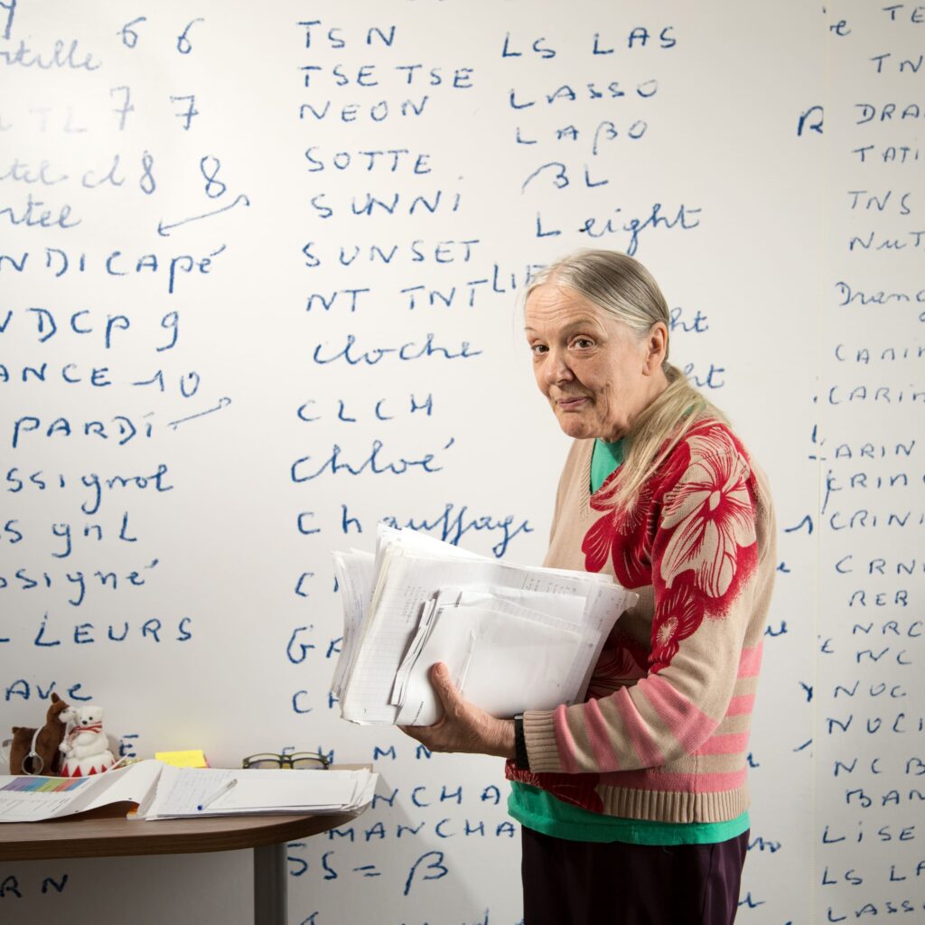 Photographie de Lionel Pralus / Light Motiv, issue de la série intitulée Désordre Ordinaire datant de 2020, représentant une femme âgée tenant une pile de feuilles devant un grand mur blanc sur lequel est inscrit du texte bleu, dans le cadre d'un projet tourné autour de mise en scène photographiques avec les patients et professionnels du SAAS.