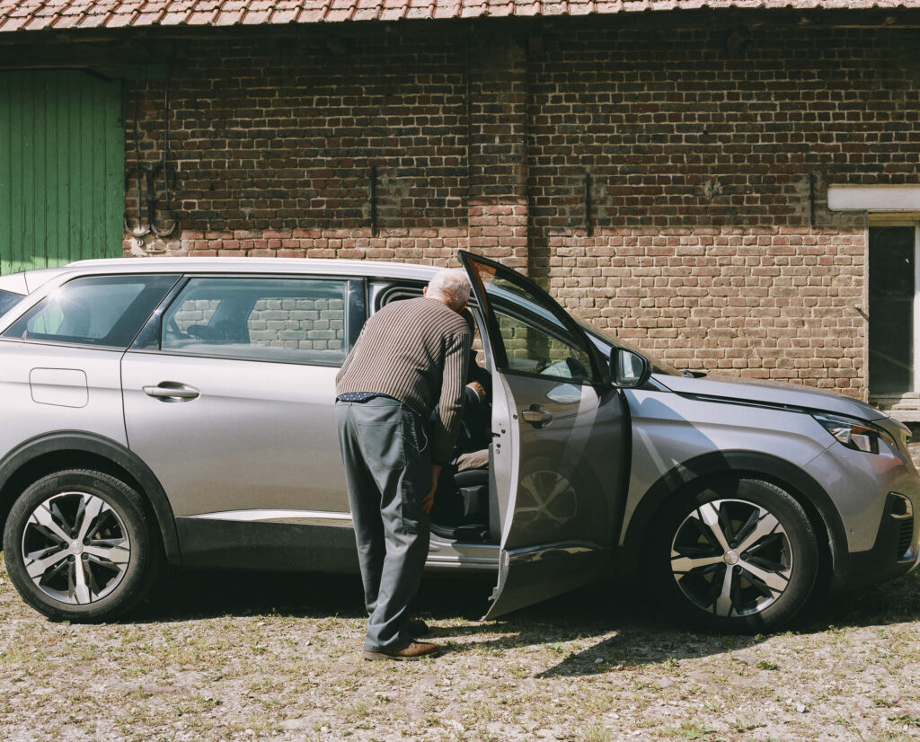Photographie de Zélie Hallosserie / Light Motiv, issue de la série intitulé Jean Marie, représentant Jean Marie dans l'entrebâillement de la portière passagère d'une voiture grise avec un bâtiment en brique rouge en arrière plan, dans l'intention de capturer les moments de vie de Jean Marie.