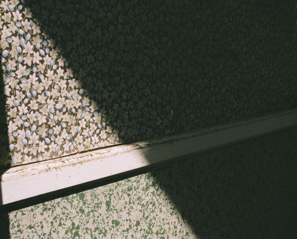 Photographie de Zélie Hallosserie / Light Motiv, issue de la série intitulé Jean Marie, représentant un mur ombragé et tapissé d'un papier peint fleuri sur lequel figure une rampe blanche en bois, dans l'intention de capturer les moments de vie de Jean Marie.