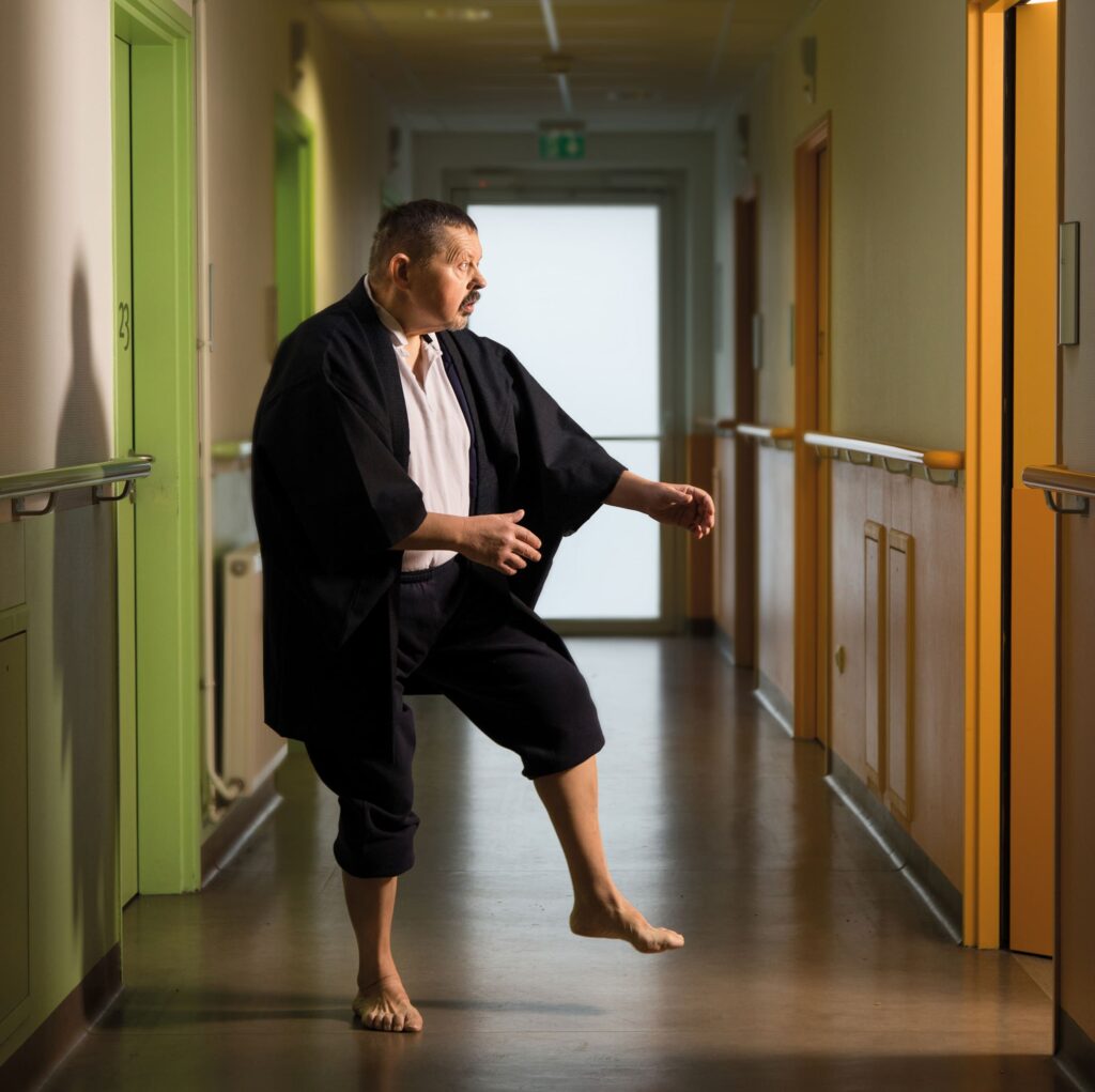 Photographie de Lionel Pralus / Light Motiv, issue de la série intitulée Désordre Ordinaire datant de 2020, représentant un homme debout sur un pied dans un couloir vide en face d'un encadrement de porte, dans le cadre d'un projet tourné autour de mise en scène photographiques avec les patients et professionnels du SAAS.