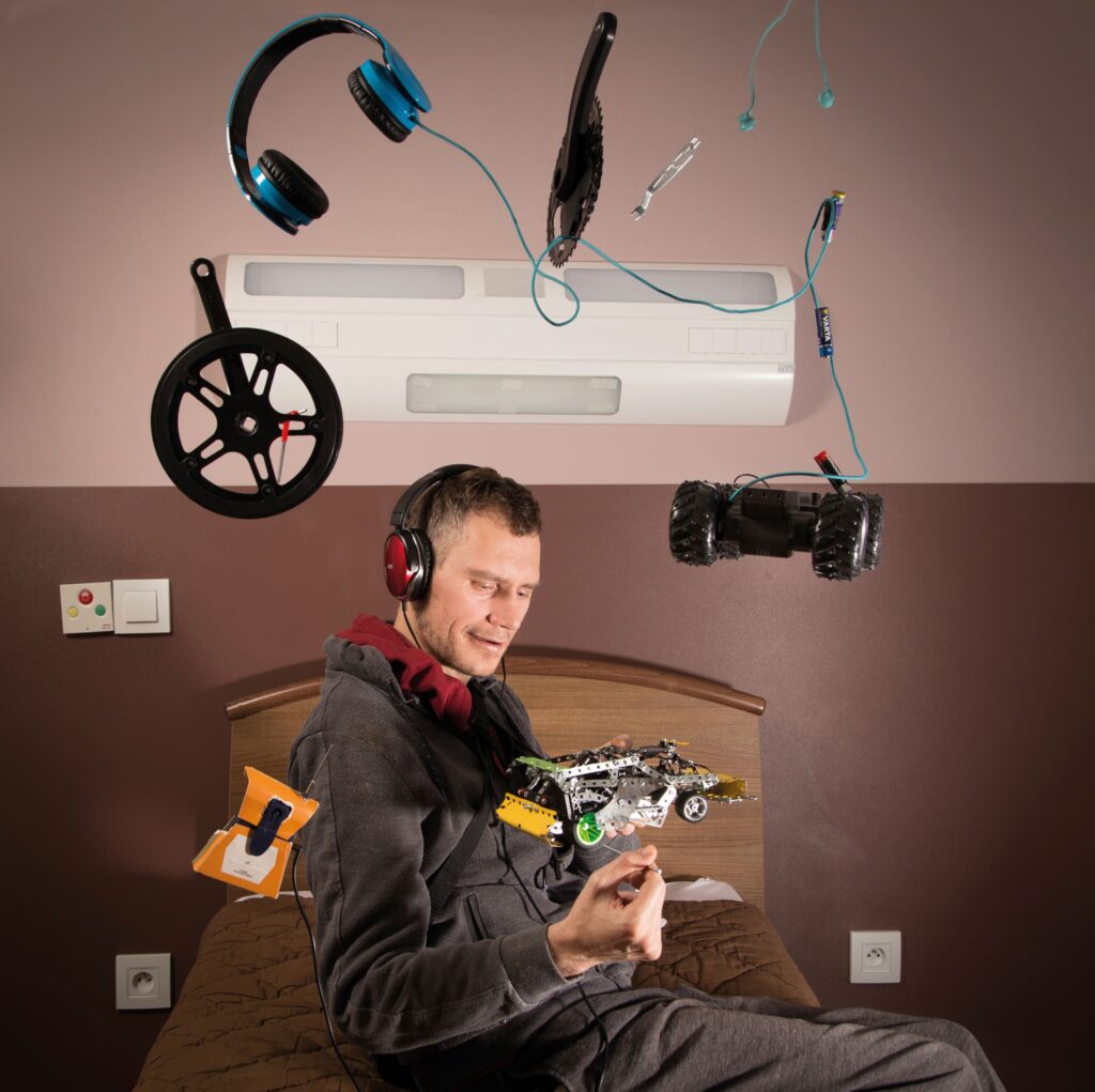 Photographie de Lionel Pralus / Light Motiv, issue de la série intitulée Désordre Ordinaire datant de 2020, représentant un homme avec un casque sur les oreilles assis sur un lit simple avec des objets qui gravite autour de lui, dans le cadre d'un projet tourné autour de mise en scène photographiques avec les patients et professionnels du SAAS.