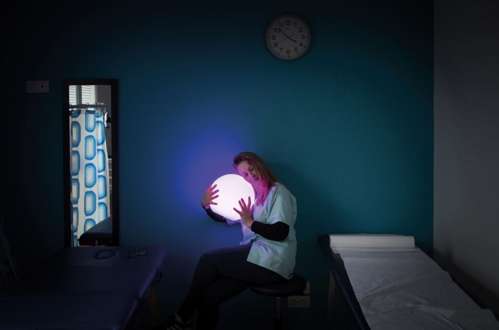 Photographie de Lionel Pralus / Light Motiv, issue de la série intitulée Désordre Ordinaire datant de 2020, représentant une femme assise sur un tabouret entre un lit et un plan de travail contre le mur tenant dans ses mains une sphère lumineuse violette, dans le cadre d'un projet tourné autour de mise en scène photographiques avec les patients et professionnels du SAAS.