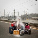 Photographie de Lionel Pralus / Light Motiv, issue de la série intitulée Désordre Ordinaire datant de 2020, représentant une femme âgée sur le quai d'une gare entourée de valises empilées avec de la poudre blanche qui la survole, dans le cadre d'un projet tourné autour de mise en scène photographiques avec les patients et professionnels du SAAS.